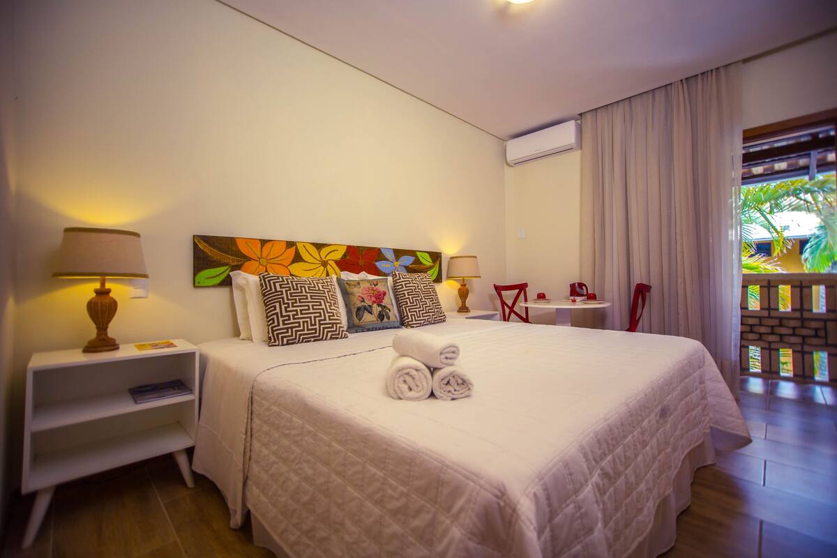 A hotel room with a large bed, two bedside tables with lamps, a colorful artwork above the bed, a red chair, and a view of greenery through a window with curtains.