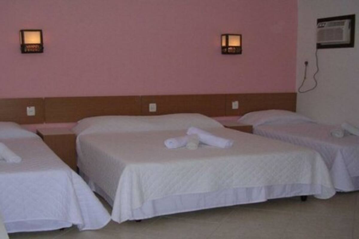 A hotel room with three single beds, each with a white bedspread and rolled towels. There are wall-mounted lamps on either side of the beds.