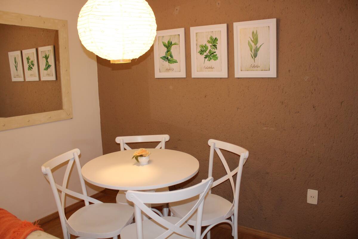 A dining area with a round table and four white chairs, a hanging light fixture, and framed botanical illustrations on the wall.