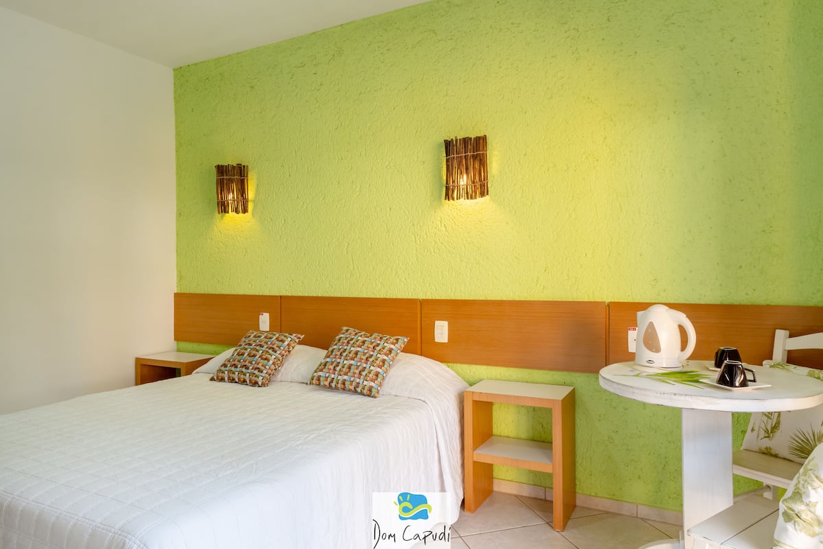 A hotel room with a bed, bedside tables, a small round table with a kettle and cups, and two wall-mounted light fixtures.
