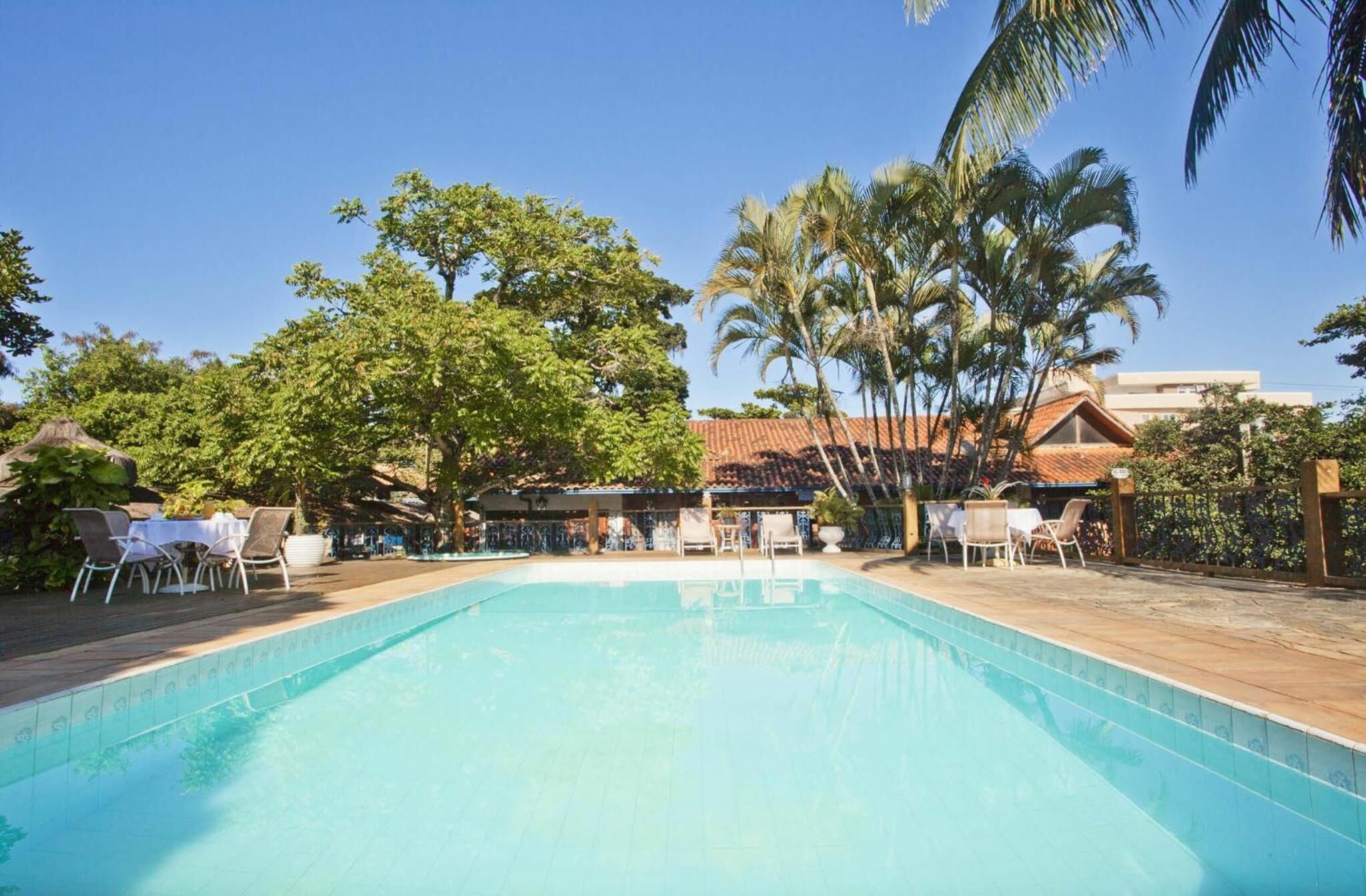 A swimming pool with lounge chairs and tables, surrounded by palm trees and a building with a tiled roof.