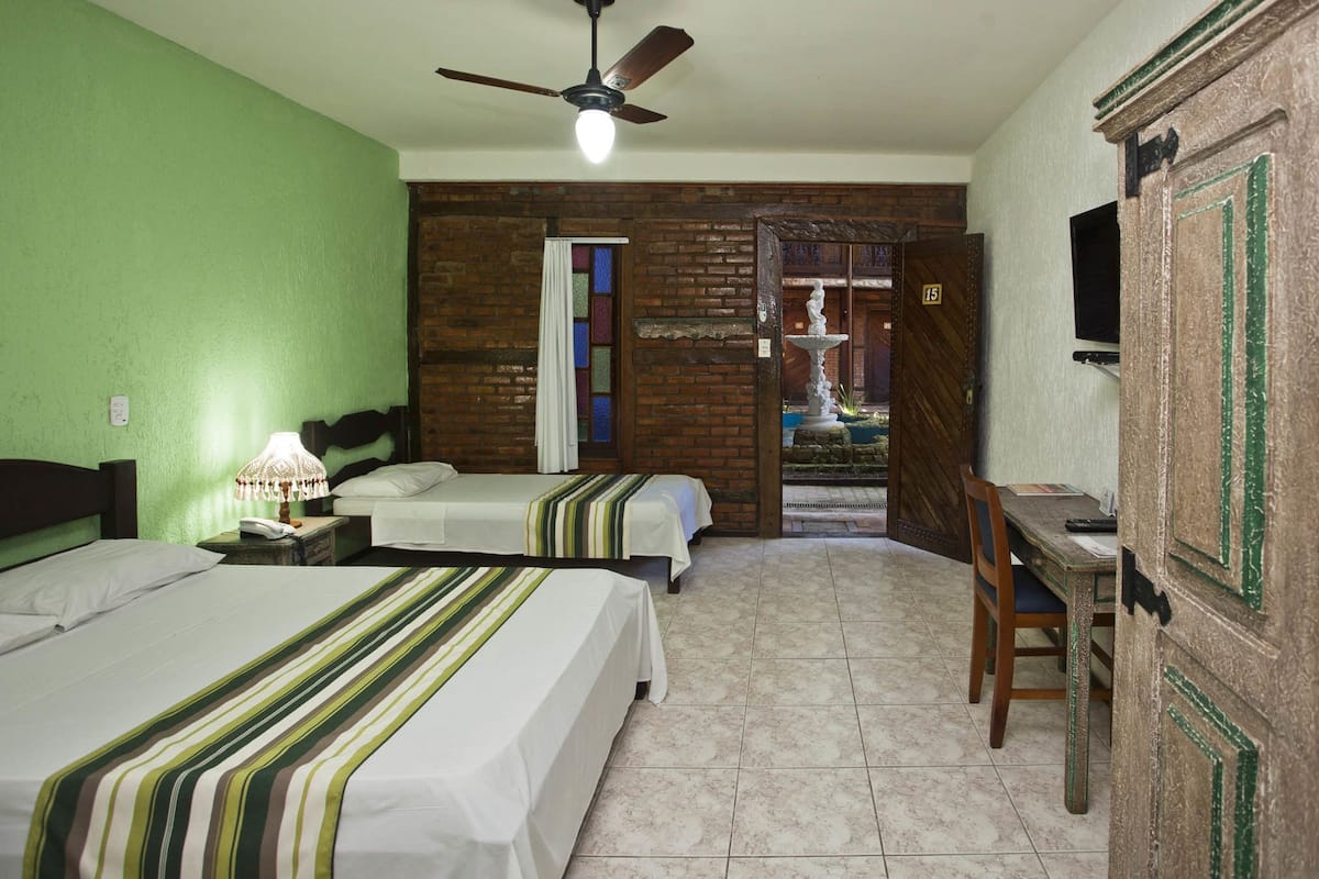 A hotel room with two beds, a desk, a chair, a ceiling fan, and a view of a courtyard.