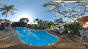 A swimming pool with lounge chairs and umbrellas, surrounded by palm trees and a stone patio. - Quari Quara BY MN Hotéis (Rio das Ostras)