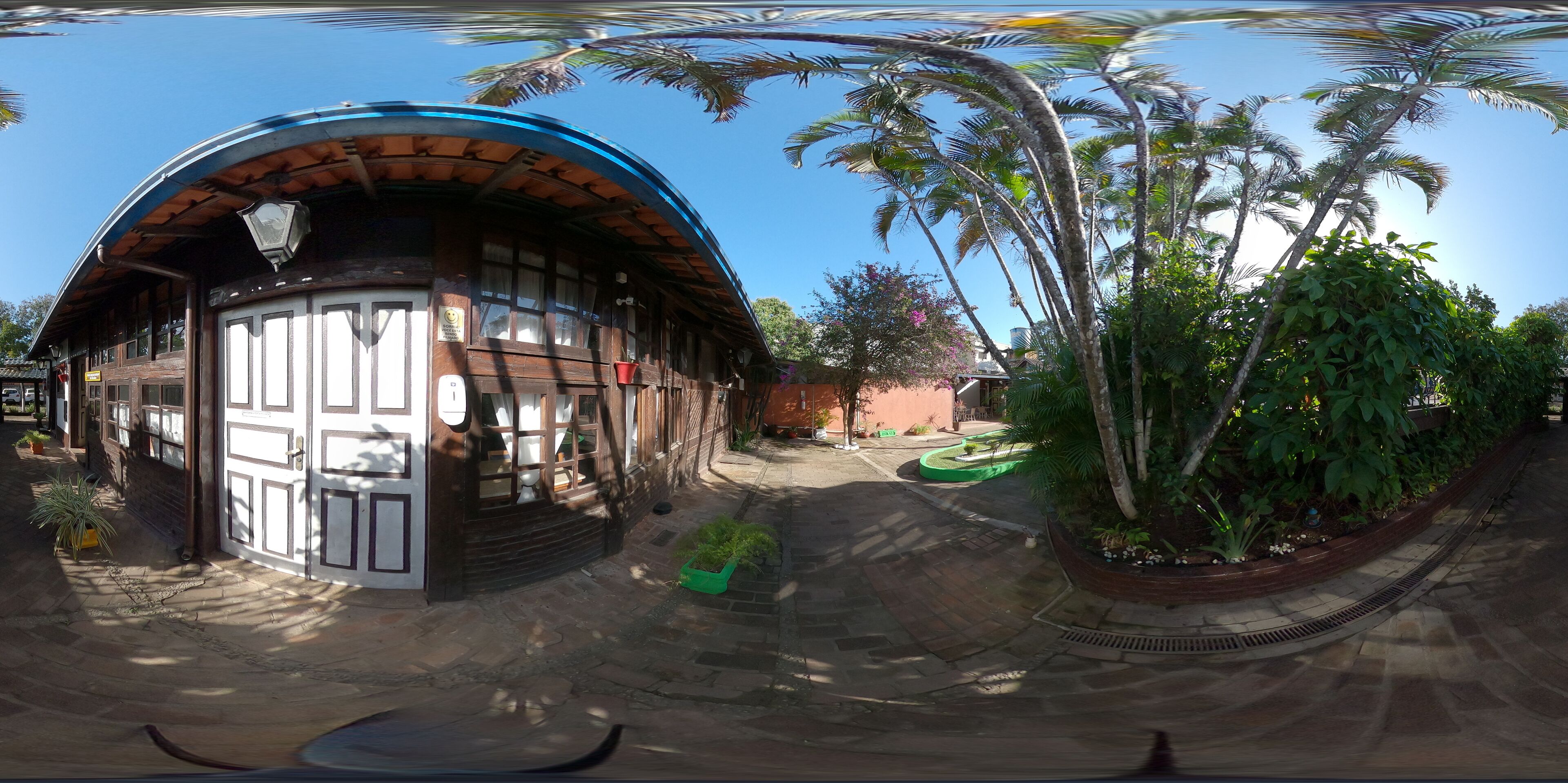 A traditional wooden building with a curved roof, a tiled entrance, and a small garden area with palm trees and potted plants.