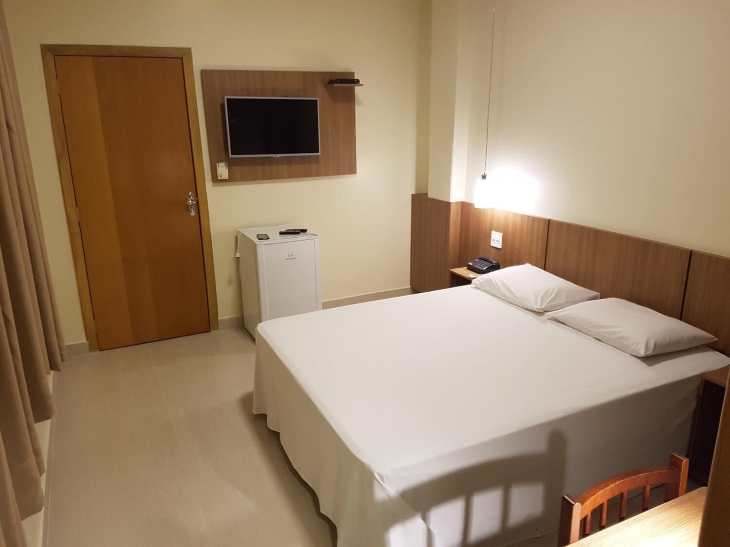 A hotel room with a bed, a desk, a TV mounted on the wall, and a small refrigerator.