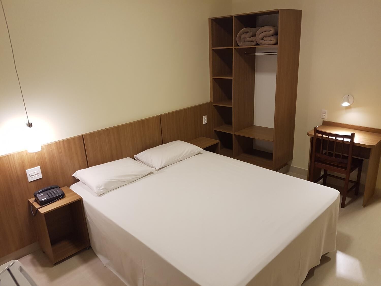 A hotel room with a single bed, a wooden wardrobe, a desk with a chair, and a bedside telephone.