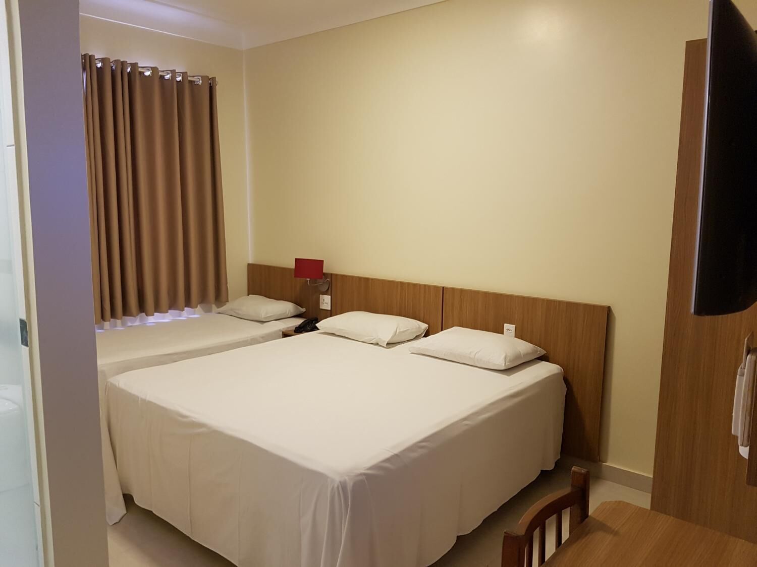 A hotel room with two beds, a wooden headboard, a bedside table with a lamp, and a TV mounted on the wall.