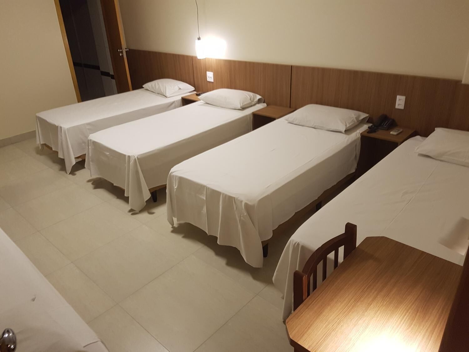 Ein Hotelzimmer mit vier Einzelbetten, die nebeneinander stehen, einem Nachttisch aus Holz mit einem Telefon und einem Holztisch.