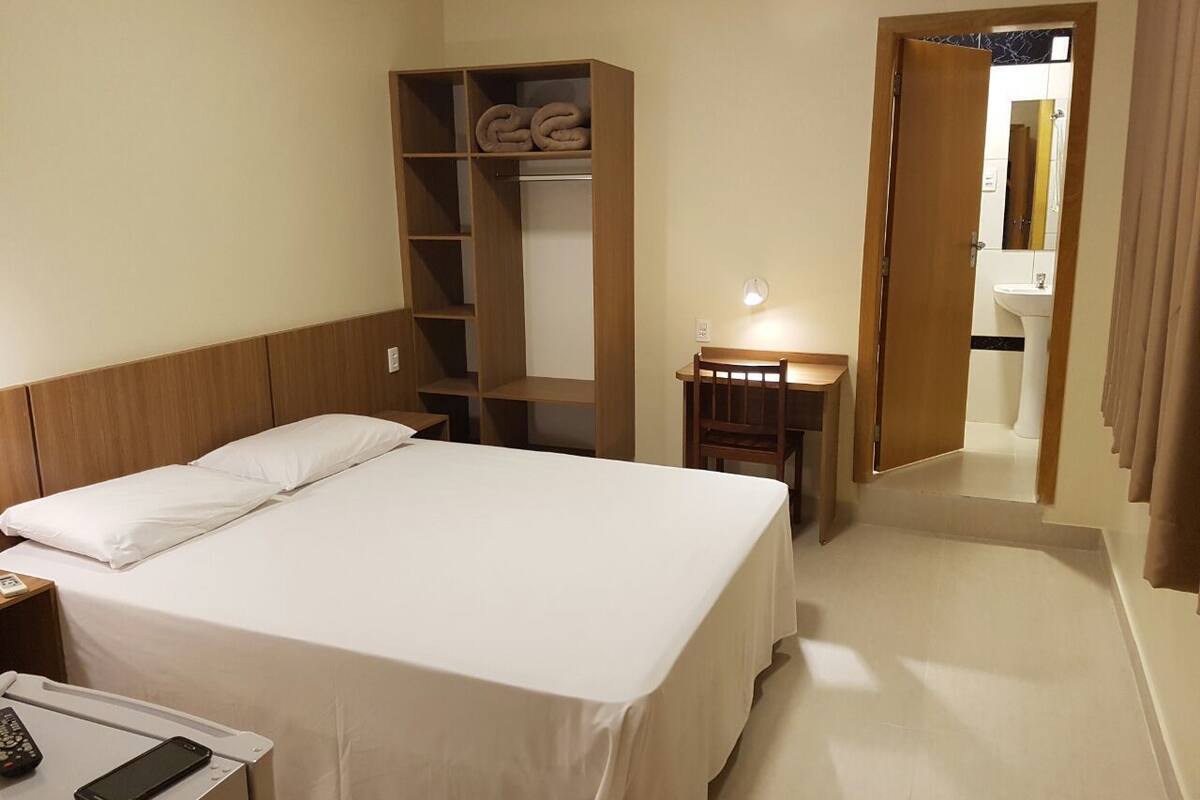 Une chambre d’hôtel avec un lit, une armoire en bois, un bureau avec une lampe et une salle de bain visible à travers une porte ouverte.