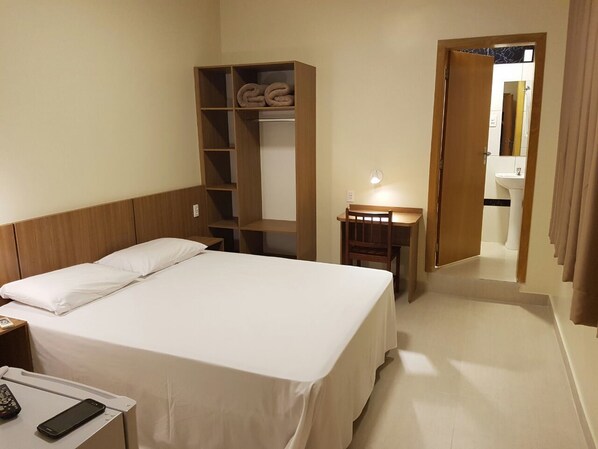 A hotel room with a bed, a wooden wardrobe, a desk with a lamp, and a bathroom visible through an open door.