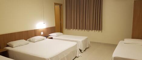 Quarto de hotel com quatro camas individuais, cabeceira de madeira, ar condicionado fixado na parede e janela com cortinas.
