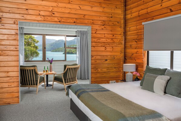 A wooden cabin room with a bed, a table with chairs, a window with a view of mountains and water, and a lamp on the nightstand. - The Portage Resort (Portage)