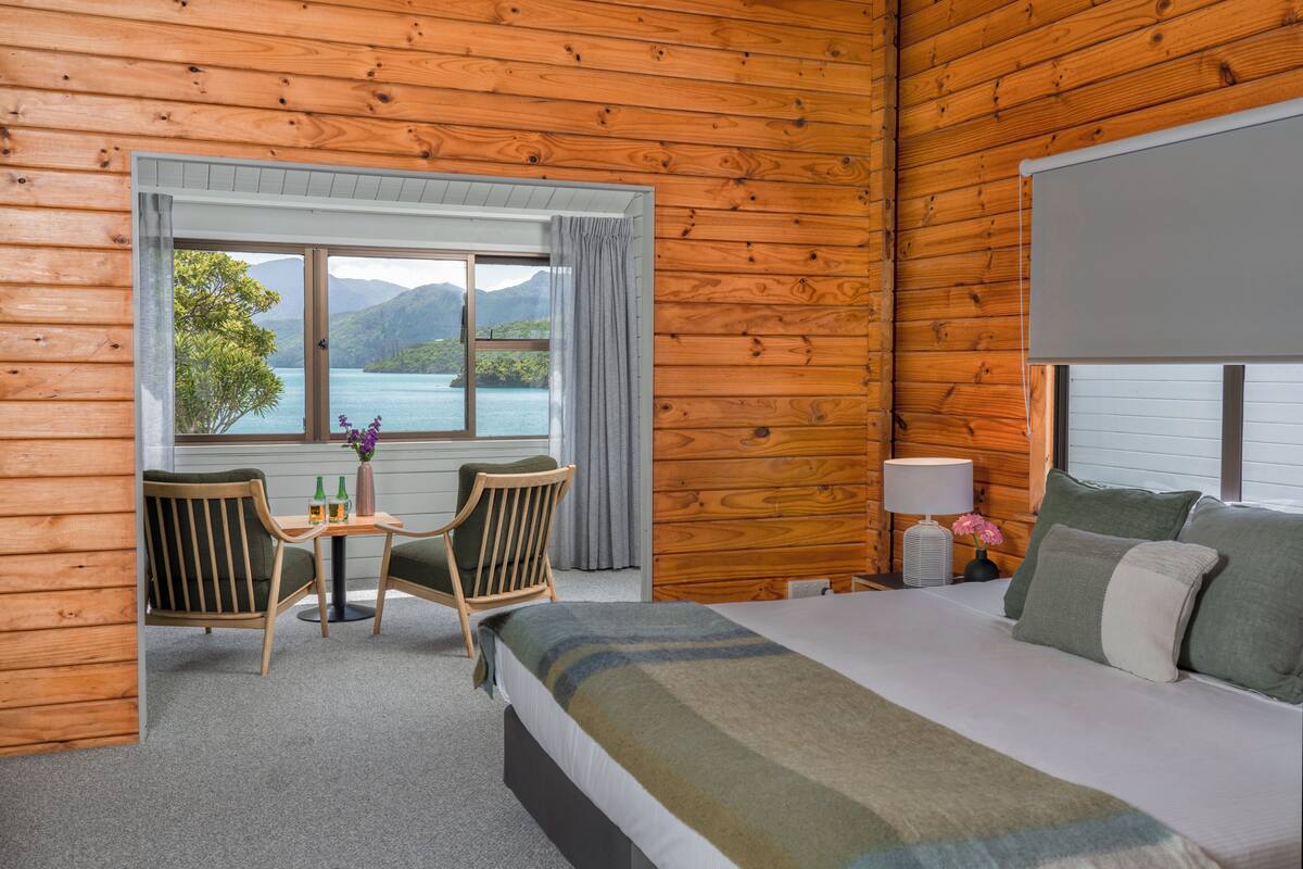 A wooden cabin room with a bed, a table with chairs, a window with a view of mountains and water, and a lamp on the nightstand.
