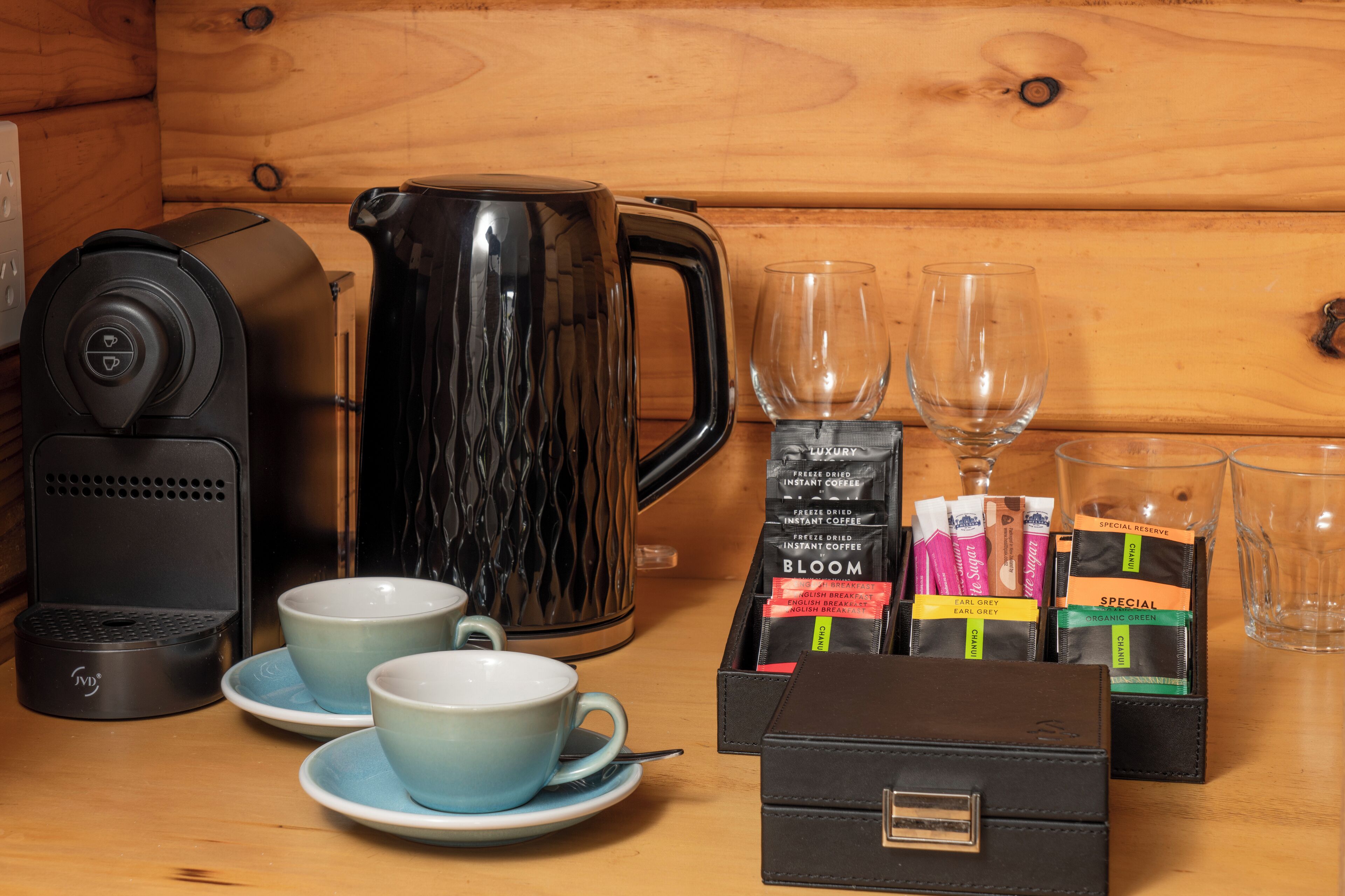 A coffee maker, two cups, a kettle, and a box of coffee pods on a wooden table.