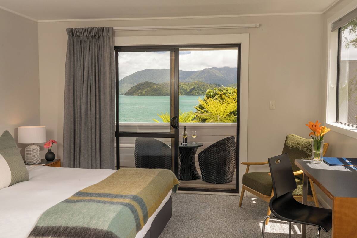 A hotel room with a bed, a desk, a chair, a table, a lamp, and a view of a lake and mountains.