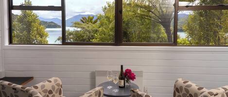 A room with a view of a lake and mountains, a round table with two chairs, and a bottle of wine.