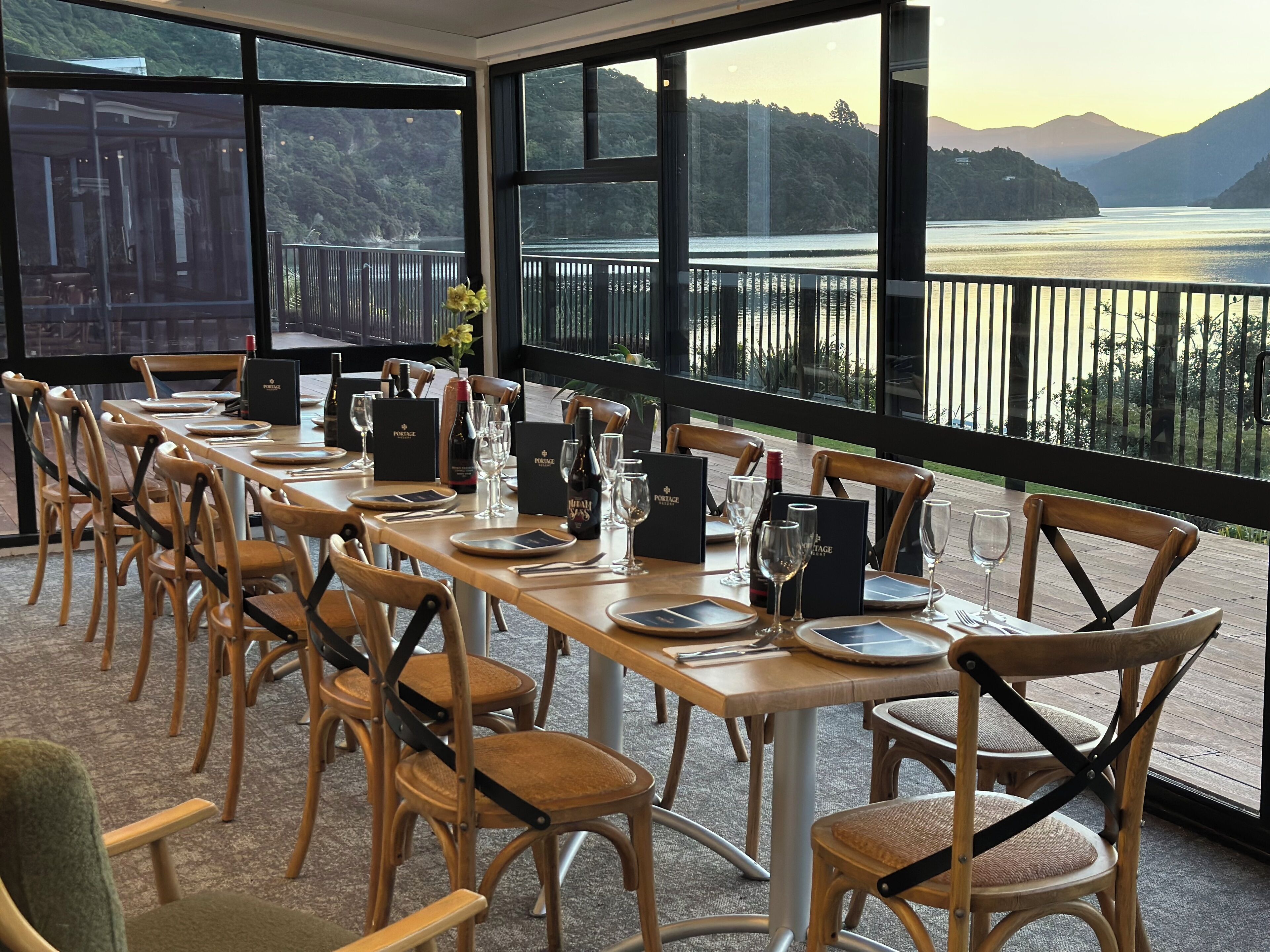 Comedor con vistas a un lago y montañas, sillas y mesas de madera dispuestas con copas de vino y platos.