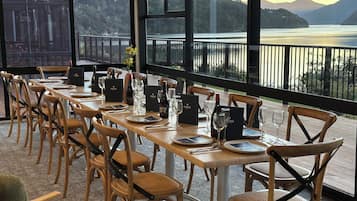 Comedor con vistas a un lago y montañas, sillas y mesas de madera dispuestas con copas de vino y platos.