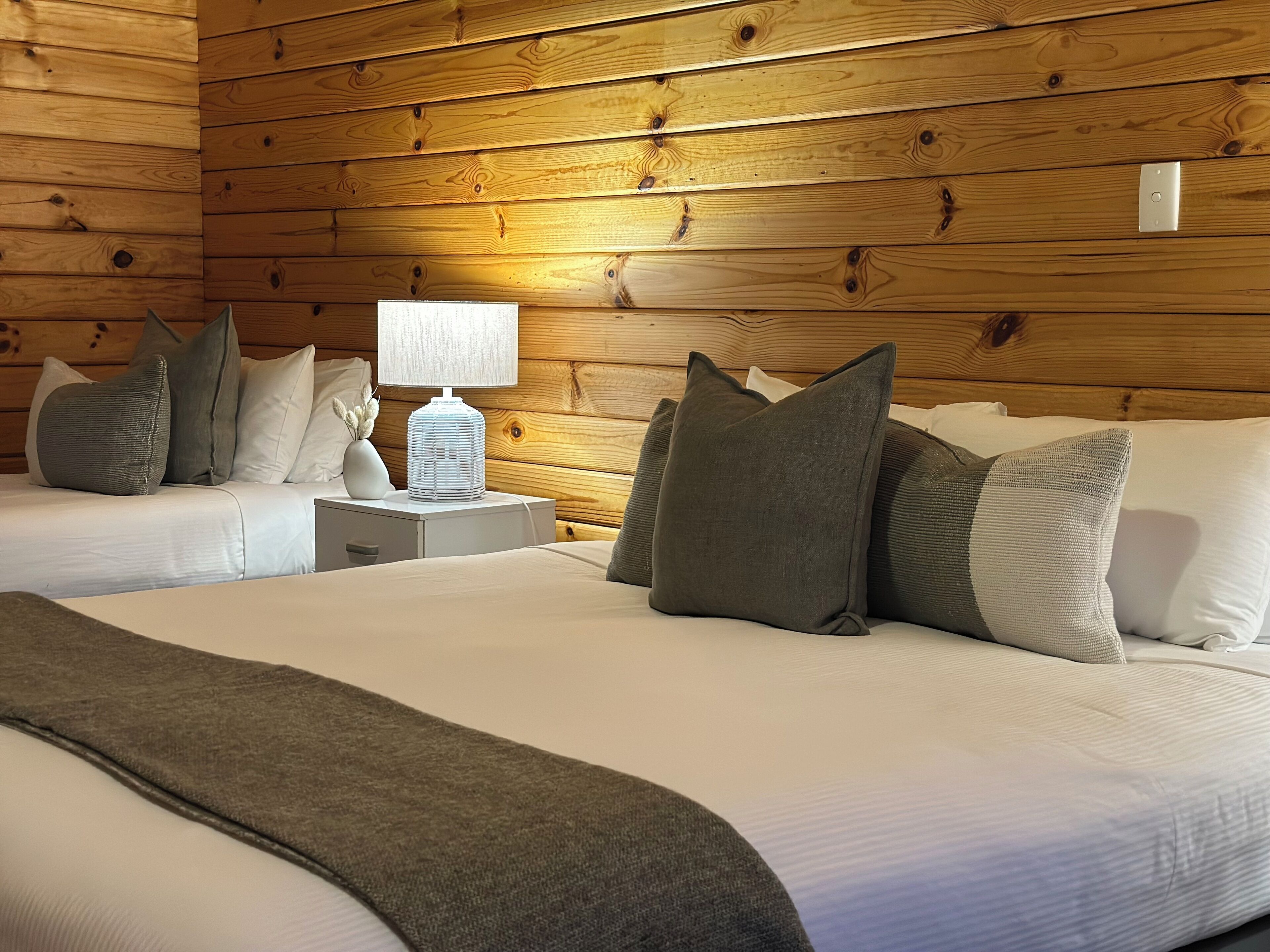 A room with two beds, wooden walls, a nightstand with a lamp, and several pillows.