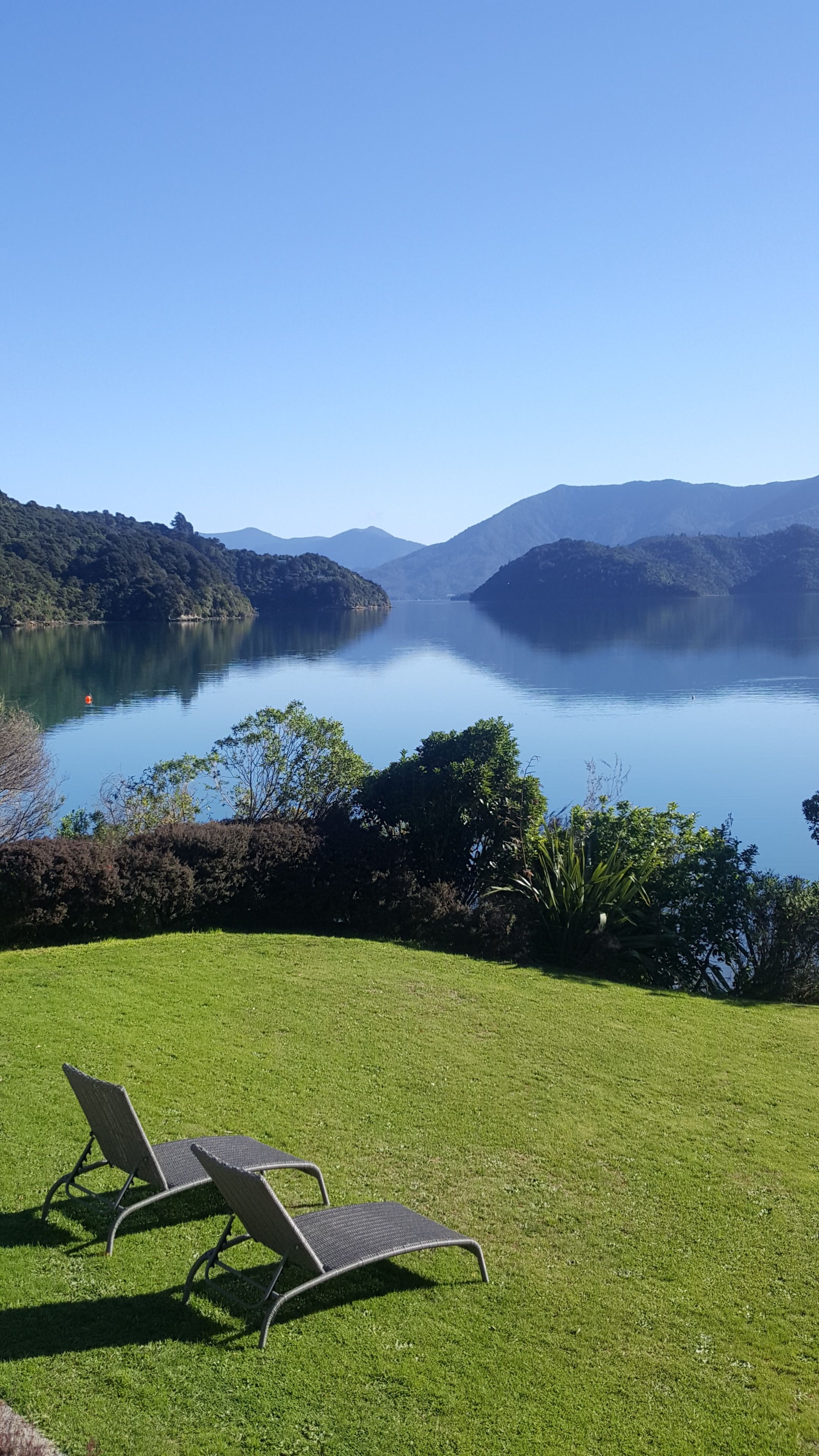 A grassy area with two lounge chairs facing a calm lake surrounded by hills.