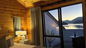 A wooden cabin room with a bed, a nightstand, a lamp, a window with a view of a lake and mountains, and a balcony with a table and chairs. - The Portage Resort (Portage)