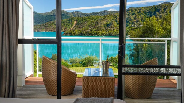 A balcony with wicker furniture, a small table with a bottle and glasses, and a view of a turquoise sea and green hills. - The Portage Resort (Portage)