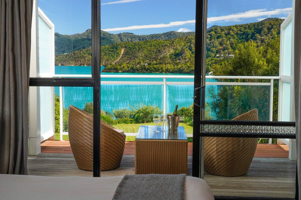 A balcony with wicker furniture, a small table with a bottle and glasses, and a view of a turquoise sea and green hills.