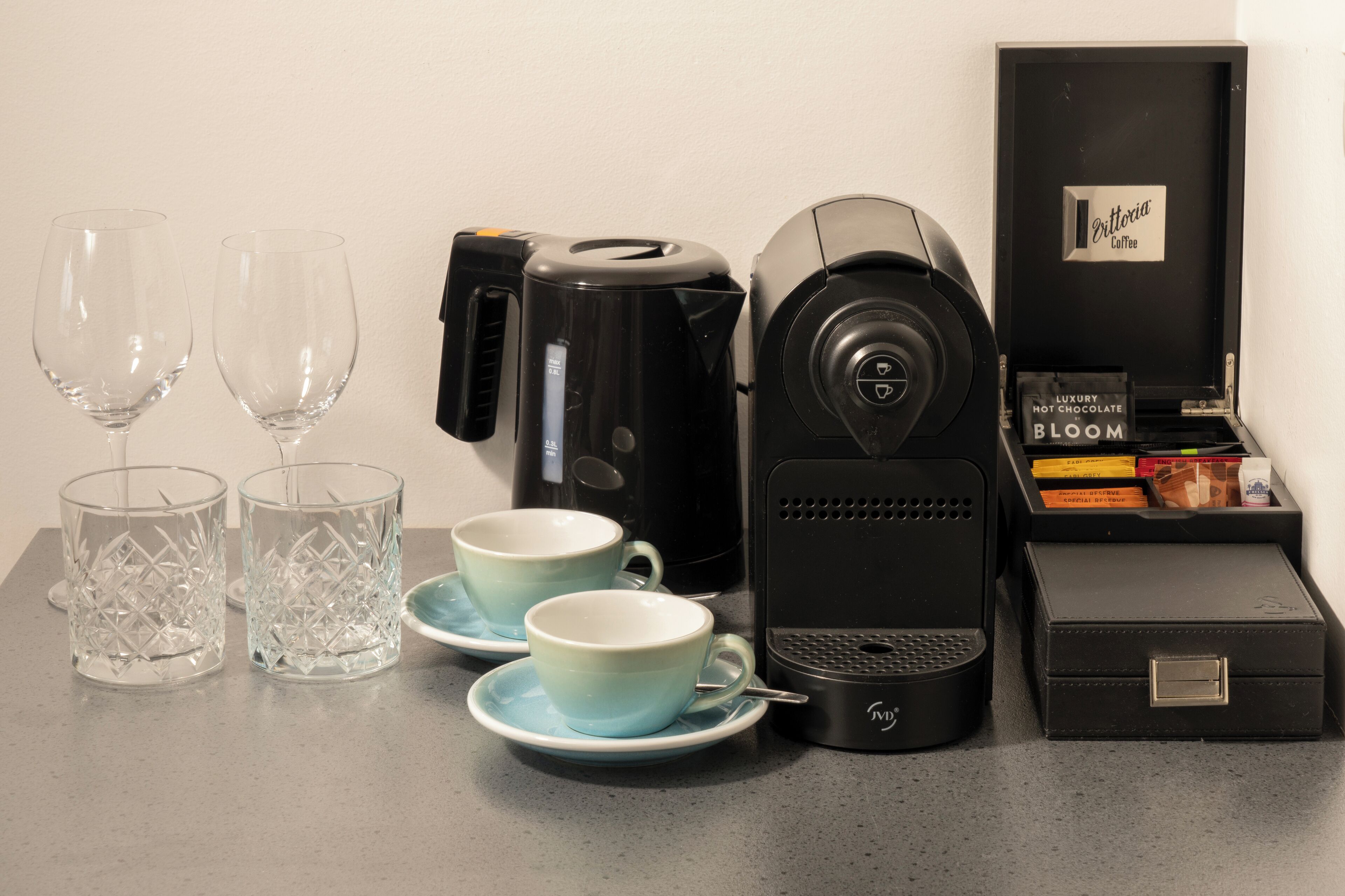 A coffee maker, two cups, two glasses, and a box of coffee beans on a table.
