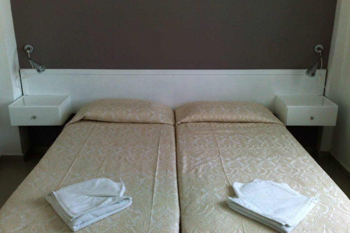 Habitación doble con dos camas, cada una con una almohada y una toalla doblada. Mesitas de noche con lámparas a ambos lados de las camas.