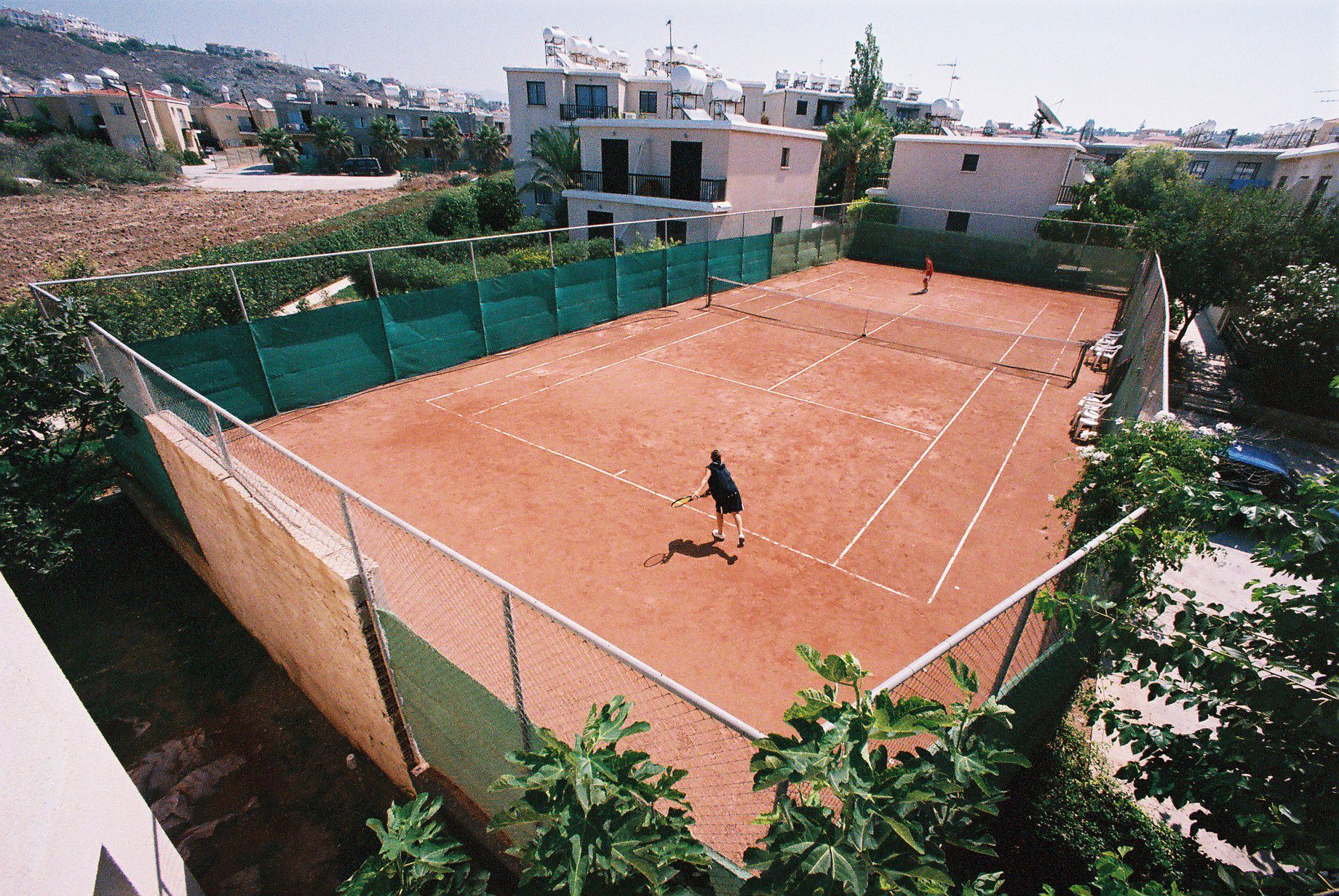 A tennis court surrounded by a green fence, with buildings in the background.