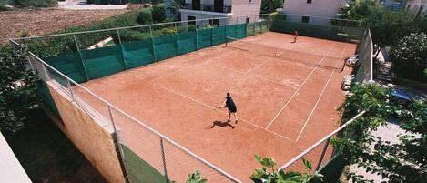 A tennis court surrounded by a green fence, with buildings in the background.