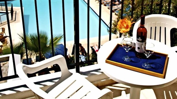 Uma varanda com cadeiras e mesa de plástico branco, com vista para uma piscina e para o litoral.