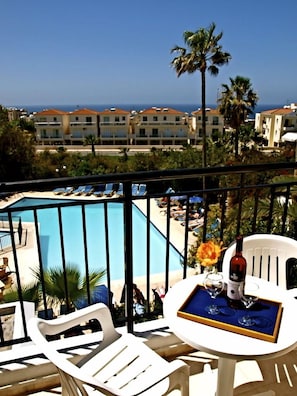A balcony with white plastic chairs and a table, overlooking a pool and a coastal view. - Hilltop Gardens (Paphos)
