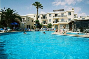 A swimming pool with lounge chairs and umbrellas, surrounded by a hotel building with balconies and a sign reading "Hilton Gardens Hotel Apartments". - Hilltop Gardens (Paphos)