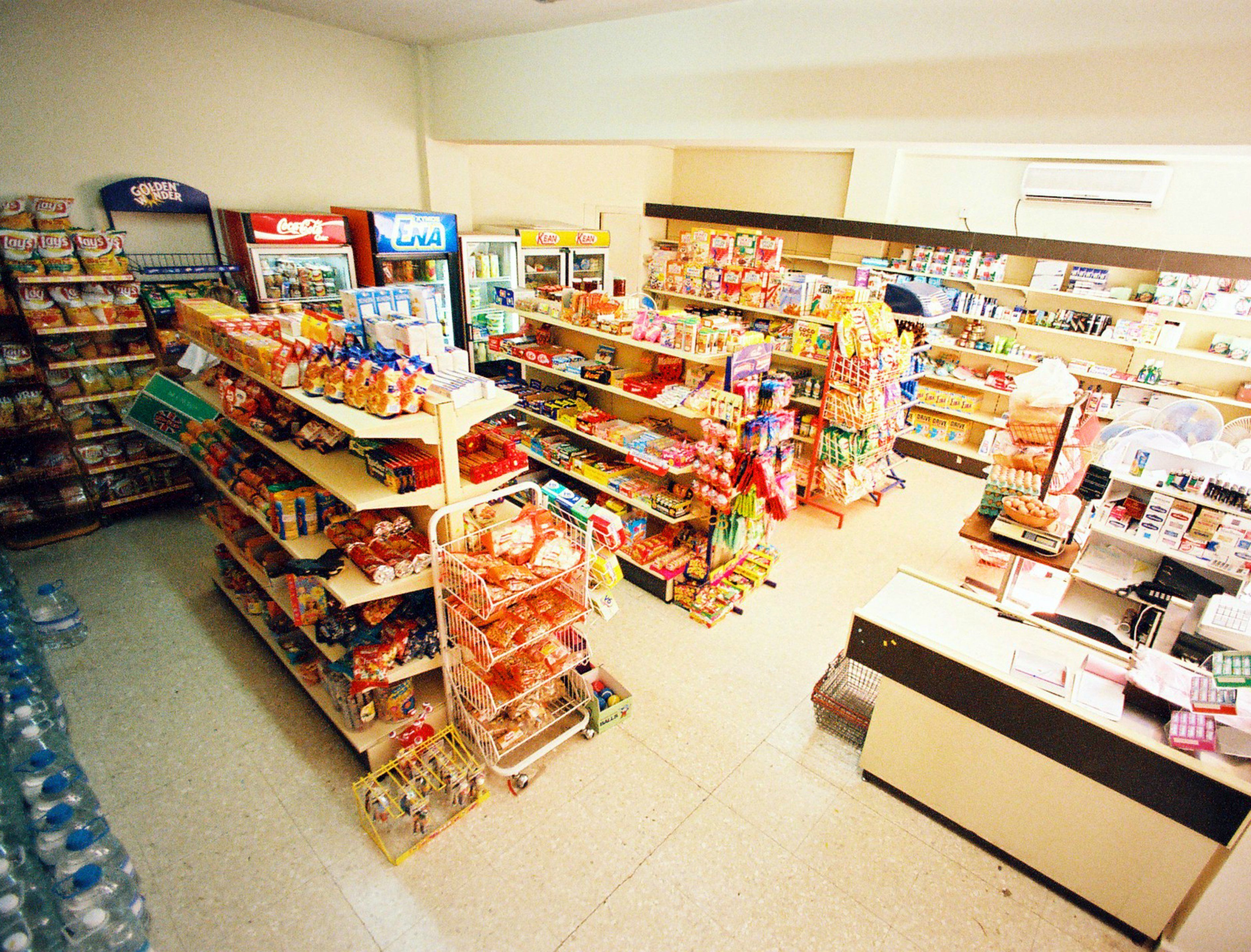 Una tienda de comestibles con estantes llenos de diversos productos, una sección refrigerada y una zona de cajas.