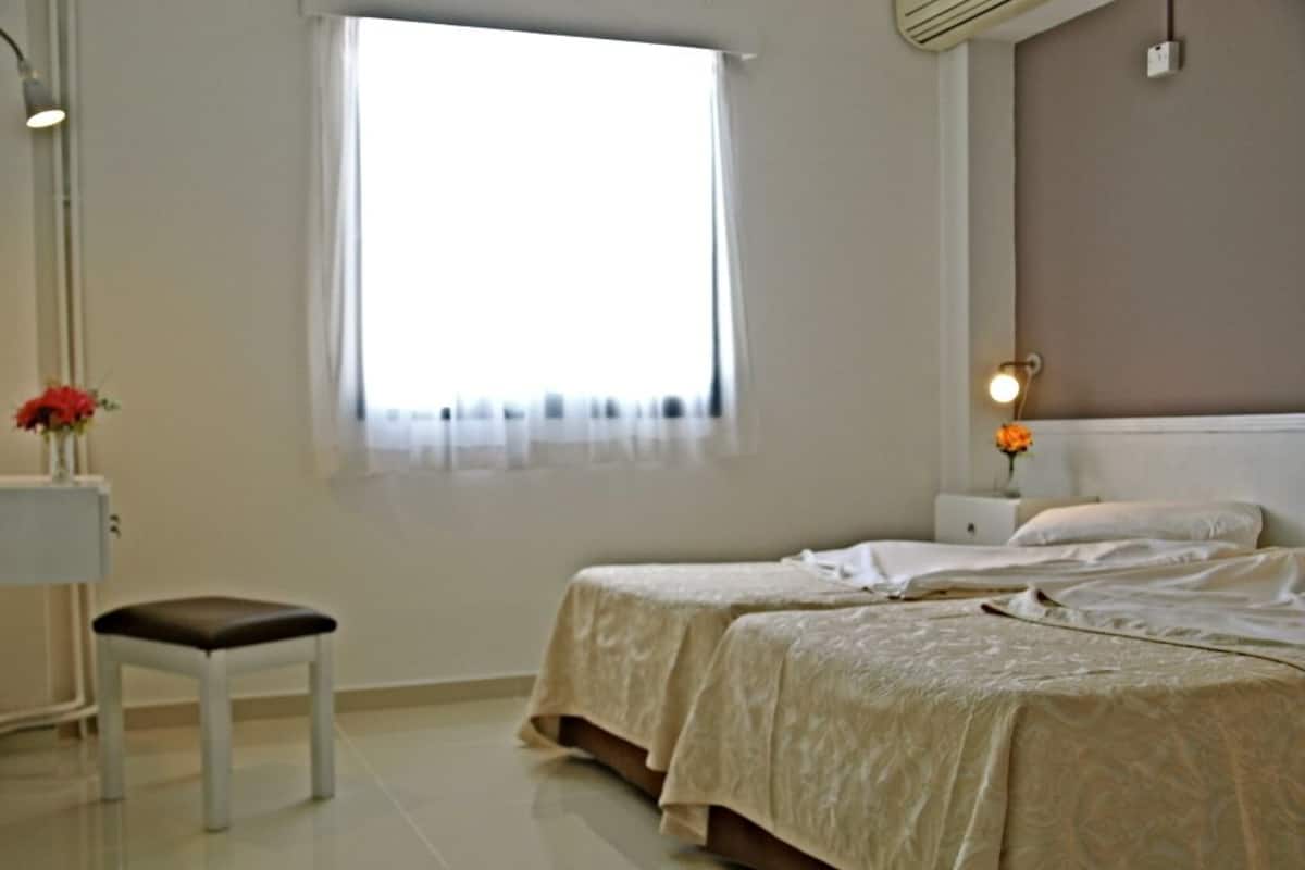 Un dormitorio con dos camas, una mesita de noche, una silla, una ventana con cortinas y una lámpara fijada a la pared.
