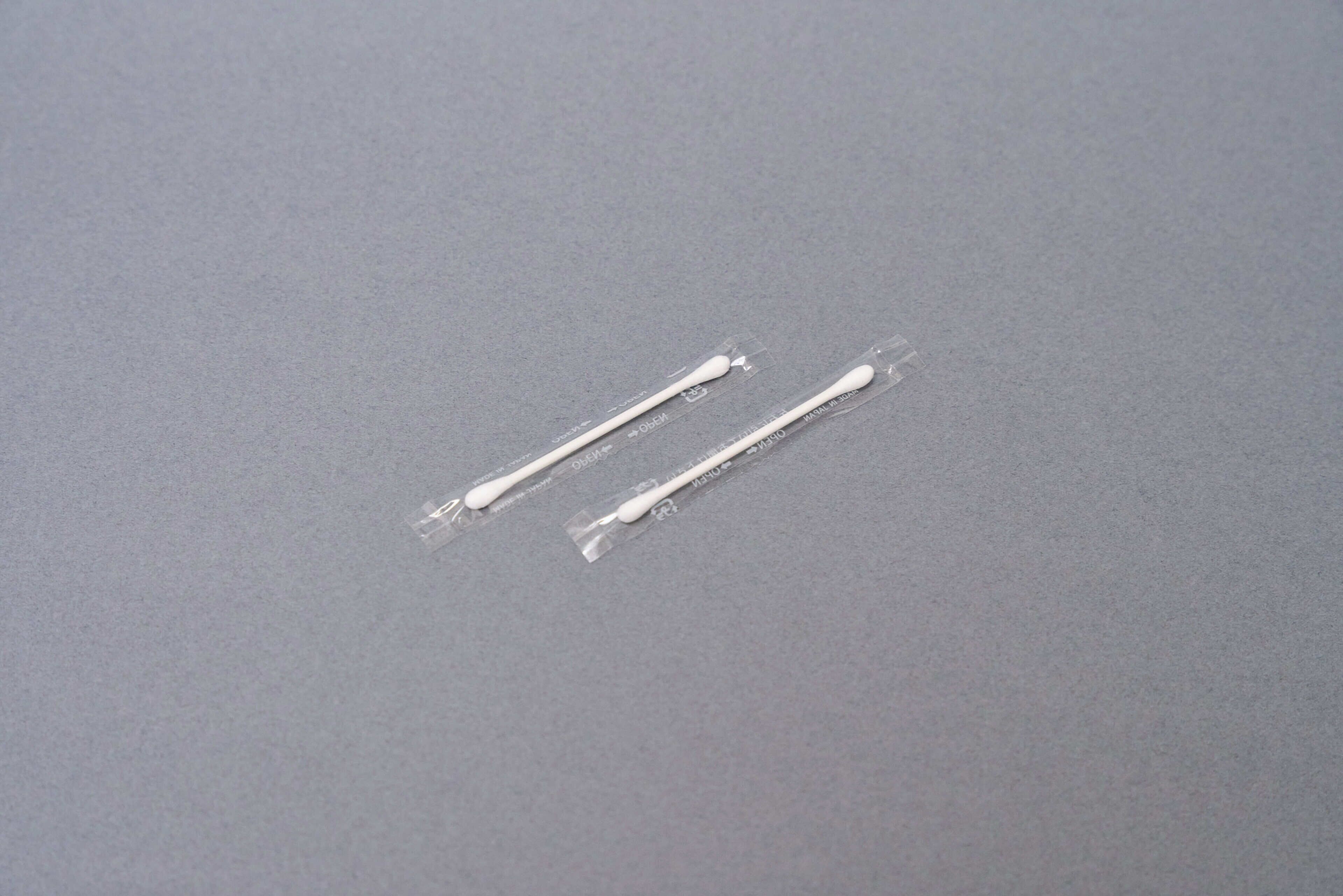 Two cotton swabs in clear plastic packaging on a grey surface.