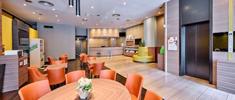 Un moderno vestíbulo de hotel con una mesa redonda central, sillas anaranjadas, un mostrador de recepción y una zona de cocina al fondo.
