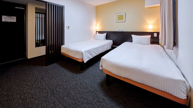A hotel room with two single beds, a dark headboard, a light-colored wall, and a small window with curtains.