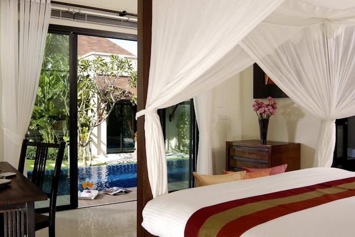 Ein Hotelzimmer mit Bett, Schreibtisch mit Laptop, Holzkommode und Blick auf einen Außenpoolbereich.