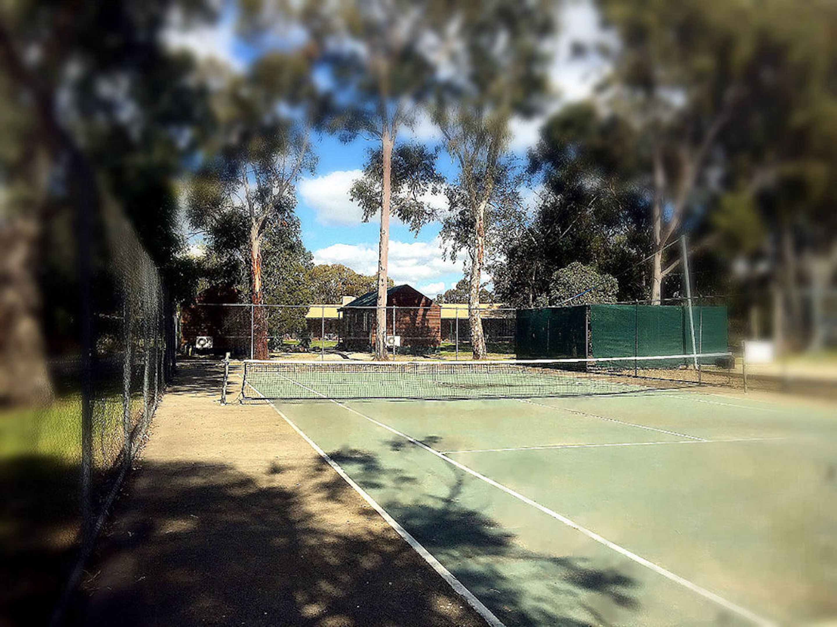 An outdoor tennis court with a net, surrounded by trees and a building in the background.