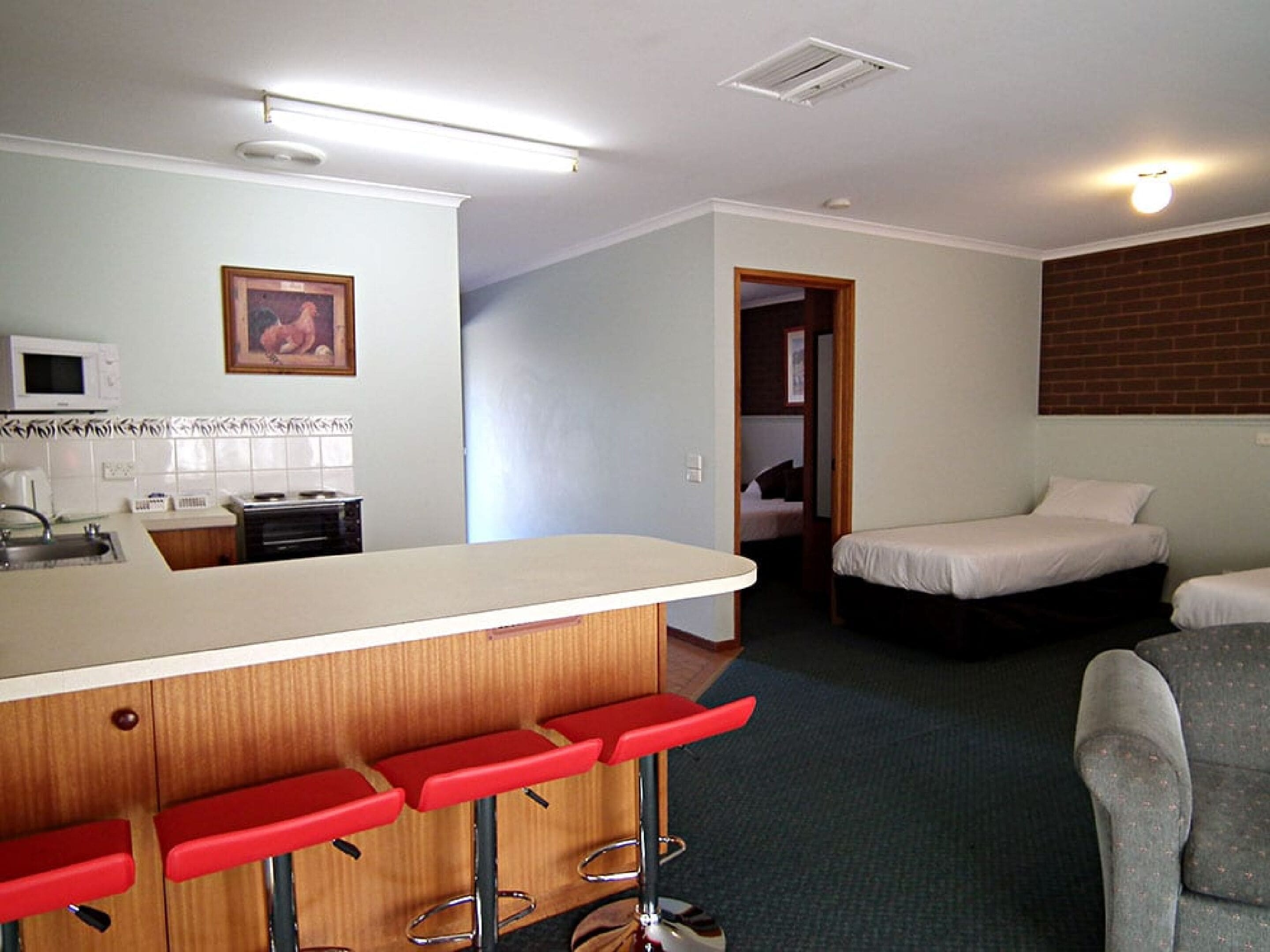 A compact hotel room with a kitchenette, a bar area with red stools, two single beds, and a grey armchair.