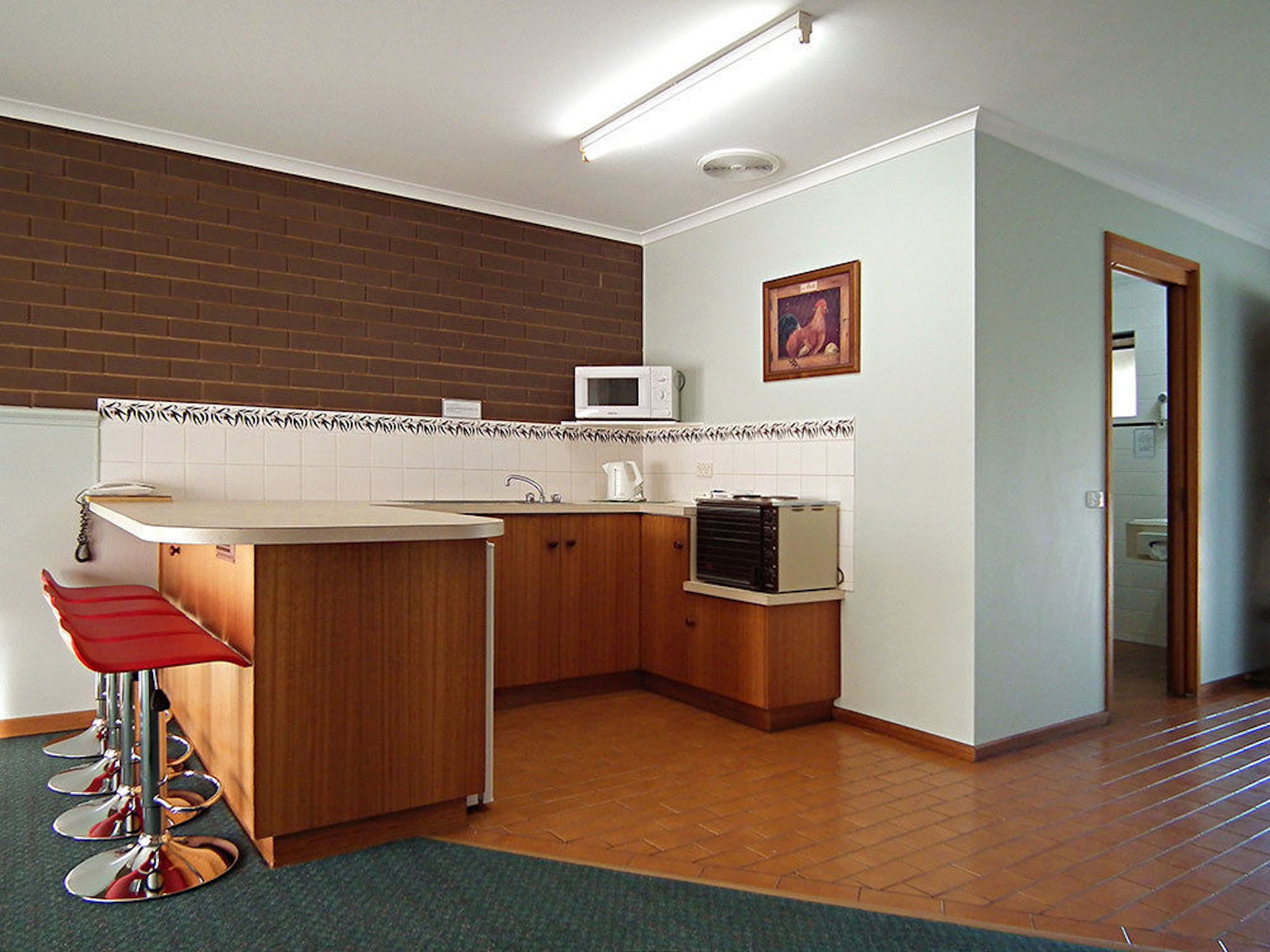 A compact kitchen with a microwave, a toaster, and a sink. There are wooden cabinets and a red bar stool.