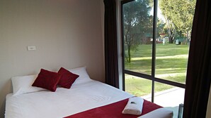 A neatly made bed with white linens and red accents, a window with a view of a grassy area and trees, and a white wall with a light switch. - Cobram Barooga Golf Resort (Barooga)