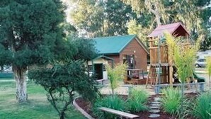 A cabin with a green roof, surrounded by a garden with a wooden deck and a small play structure. - Cobram Barooga Golf Resort (Barooga)