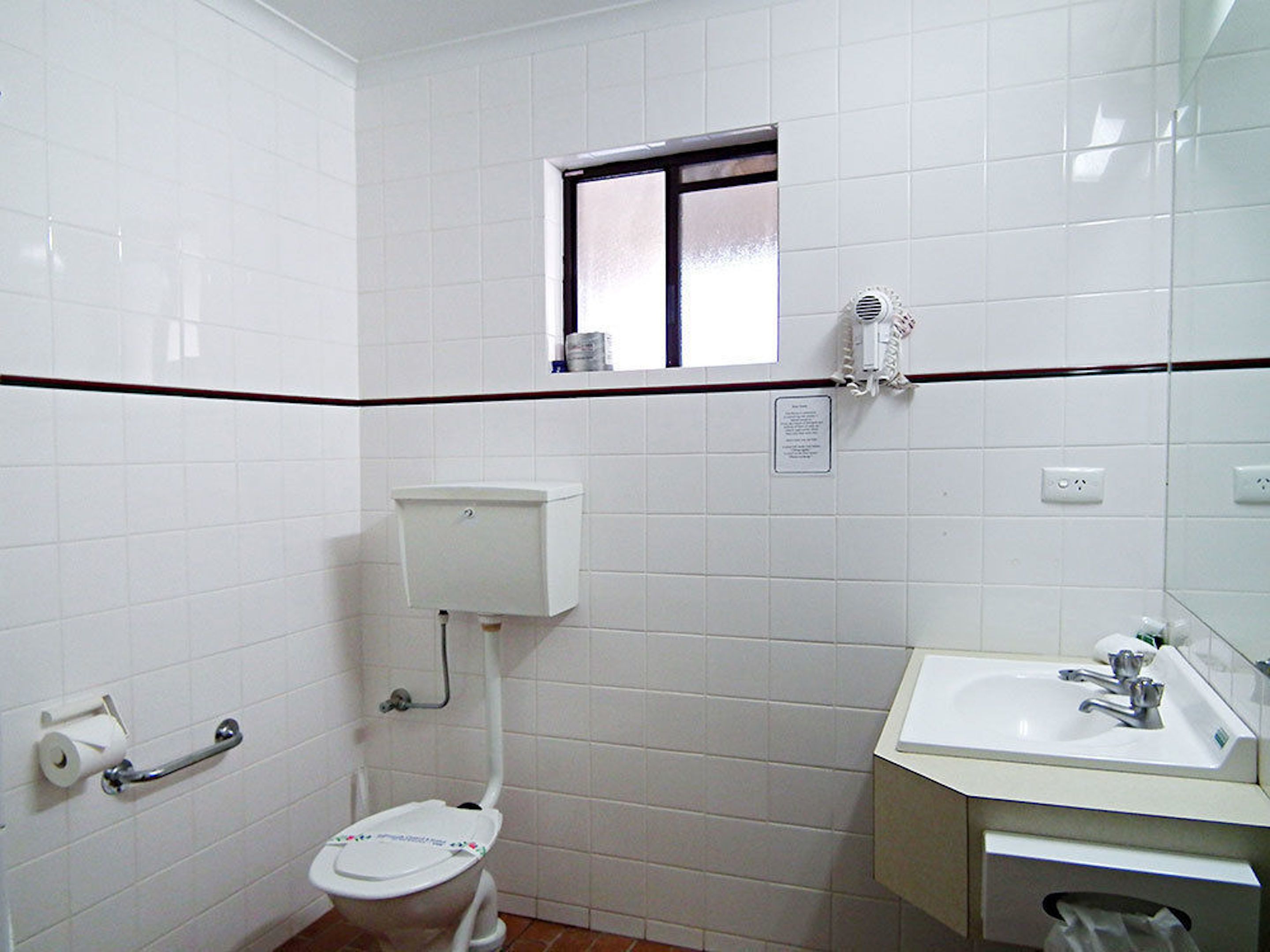 Un baño pequeño con paredes de azulejos blancos, un inodoro, un lavamanos y un ventanal sobre el inodoro.