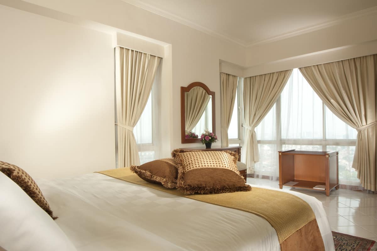 Ein Hotelzimmer mit einem großen Bett, einem Holfnachtkästchen, einem Spiegel und Blick auf die Stadt durch große Fenster.