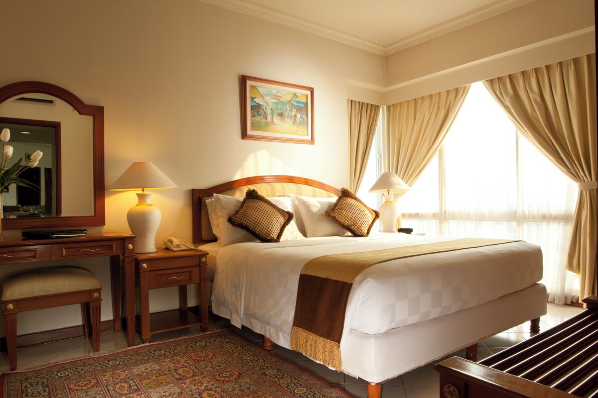 Une chambre d’hôtel avec un grand lit, un bureau avec une lampe, une chaise, une table de chevet avec une lampe, un tableau au mur et une fenêtre avec des rideaux.