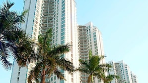 Un edificio residencial alto con varios balcones, palmeras frente a él y un pequeño edificio con techo de tejas.