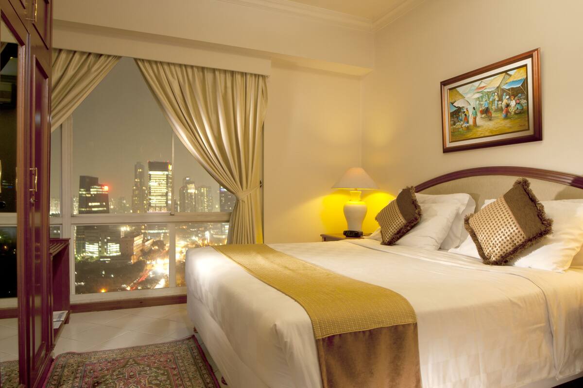 Ein Hotelzimmer mit einem großen Bett, Blick auf eine Stadtlandschaft durch ein Fenster und einem Bild an der Wand.